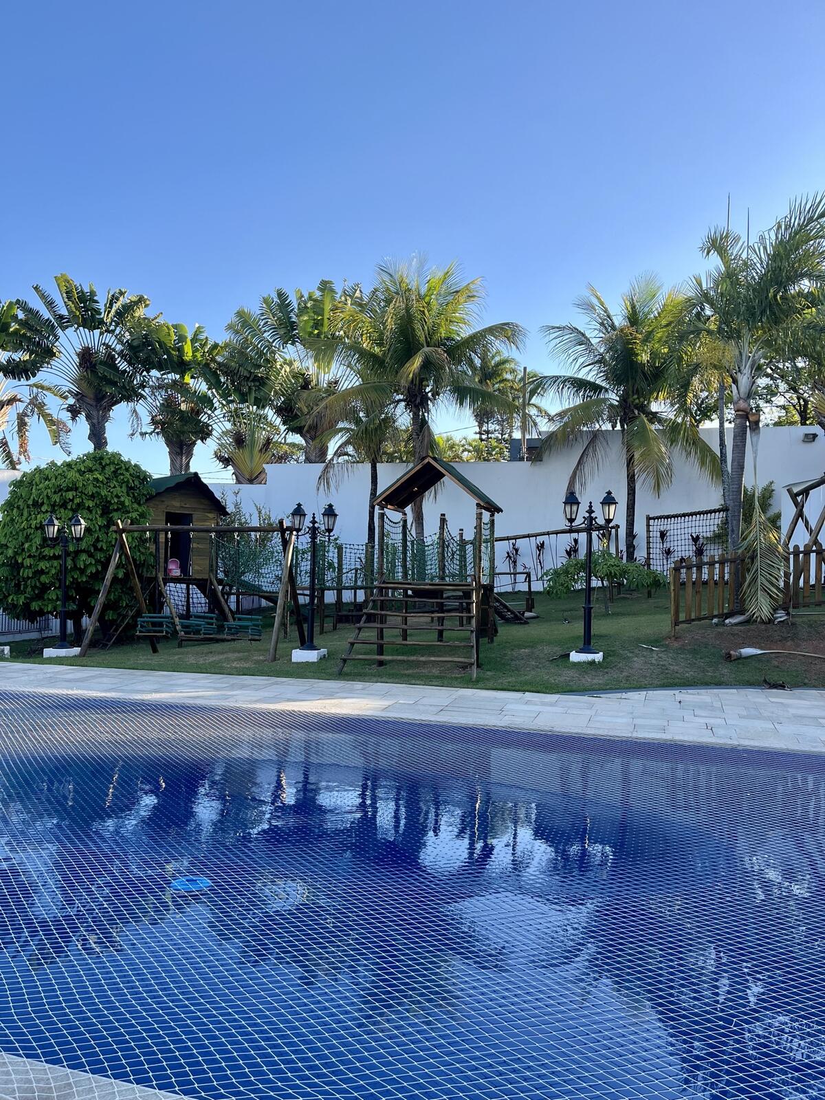 Piscina cercada de coqueiros com playground ao fundo, no Espaço Martinica